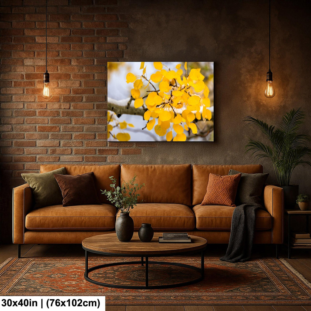 a cozy living room with a brown leather sofa, a wooden coffee table, and a brick wall backdrop. A large framed print of a yellow tree with orange leaves hangs above the sofa, adding a touch of nature to the space. The room is illuminated by two hanging light fixtures, creating a warm and inviting atmosphere.