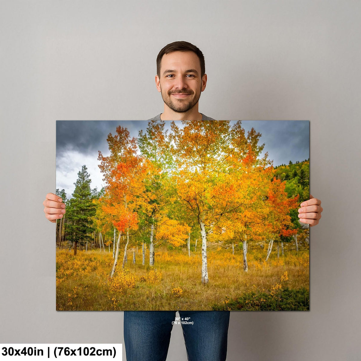A man is holding up a large canvas print of a vibrant autumn landscape featuring a forest of trees with orange and yellow foliage.