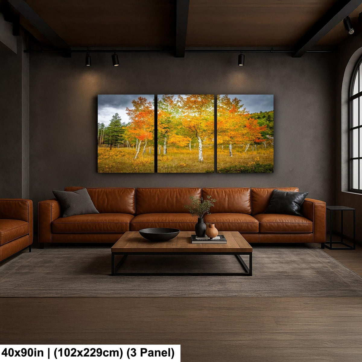 a cozy living room with a large, three-panel landscape painting hanging on the wall above a brown leather sofa. The painting features a scenic view of a forest with vibrant autumn foliage. The room has a modern, industrial design with exposed beams and a wooden floor.