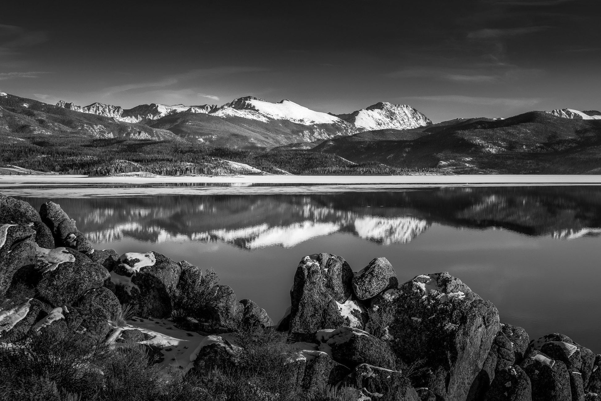 black and white photo of Grand Lake Colorado in January 2024. Mountains with snowy peaks reflecting in the water with rocks in the foreground.Available in canvas or paper prints.