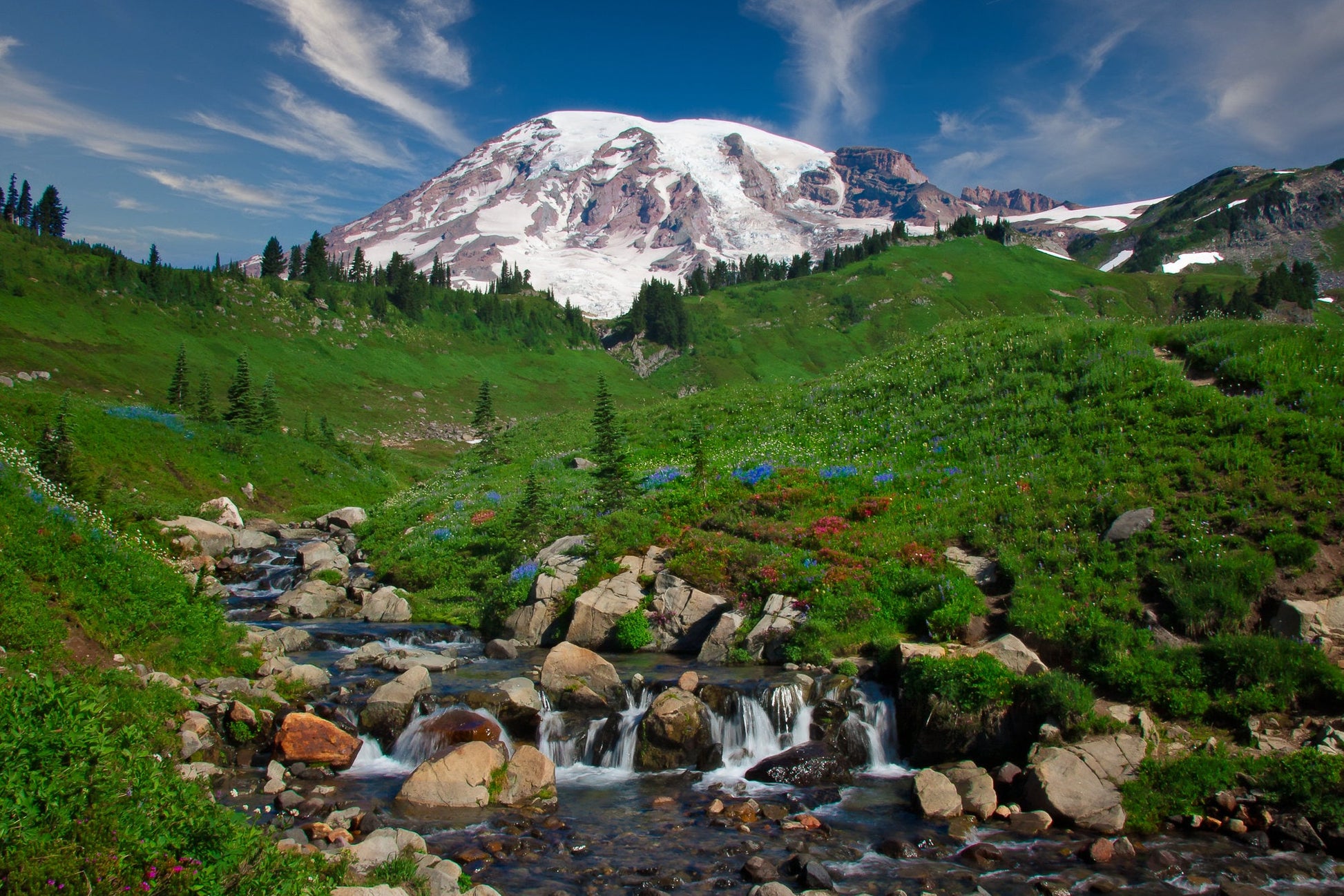 Beautiful mountain landscape, with snowy mountain peak soaring above green slopes and a creek babbling over rocks and wildflowers. Available in canvas and fine art print in both horizontal and vertical orientation.