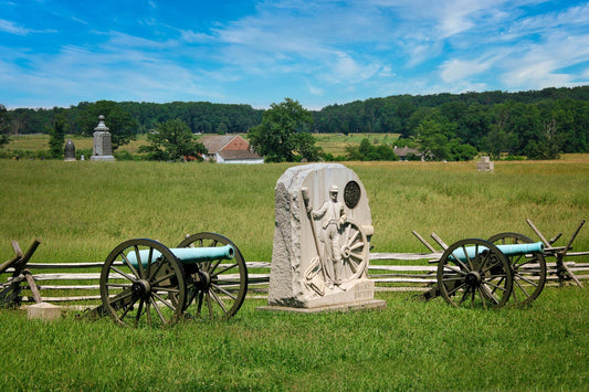 Civil war cannons and monument in front of split rail fence bordering field of battle of Gettysburg.